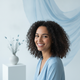 A smiling woman with light brown skin and dark curly hair, wearing a sky blue top, against a bright white and slate blue back