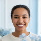 A smiling young woman with warm eyes, looking at the viewer. Her expression is friendly and trustworthy, against a soft, brig
